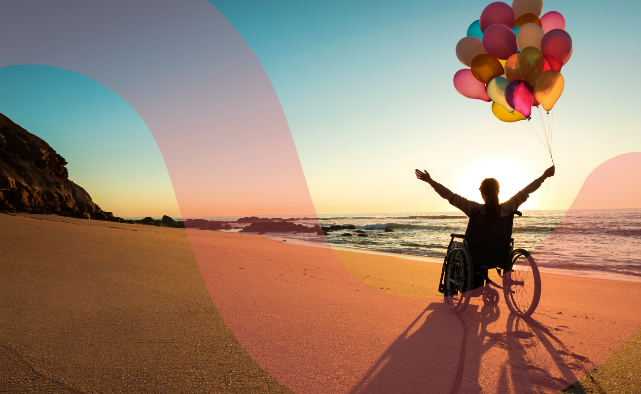 A photograph of an person on a beach with arms raised. They are holding balloons and celebrating a sunset.