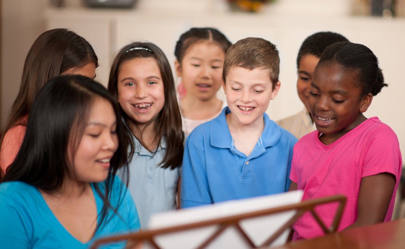 A photograph of a group of children singing. An adult is sitting at a piano and has a sheet of music.